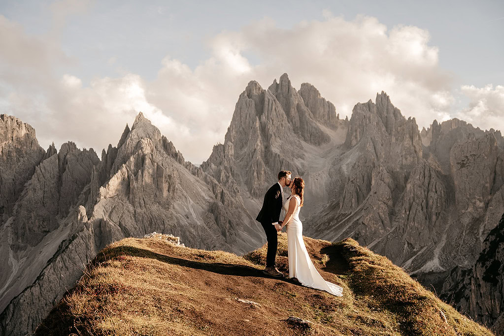Couple kissing on mountain with rocky peaks backdrop.