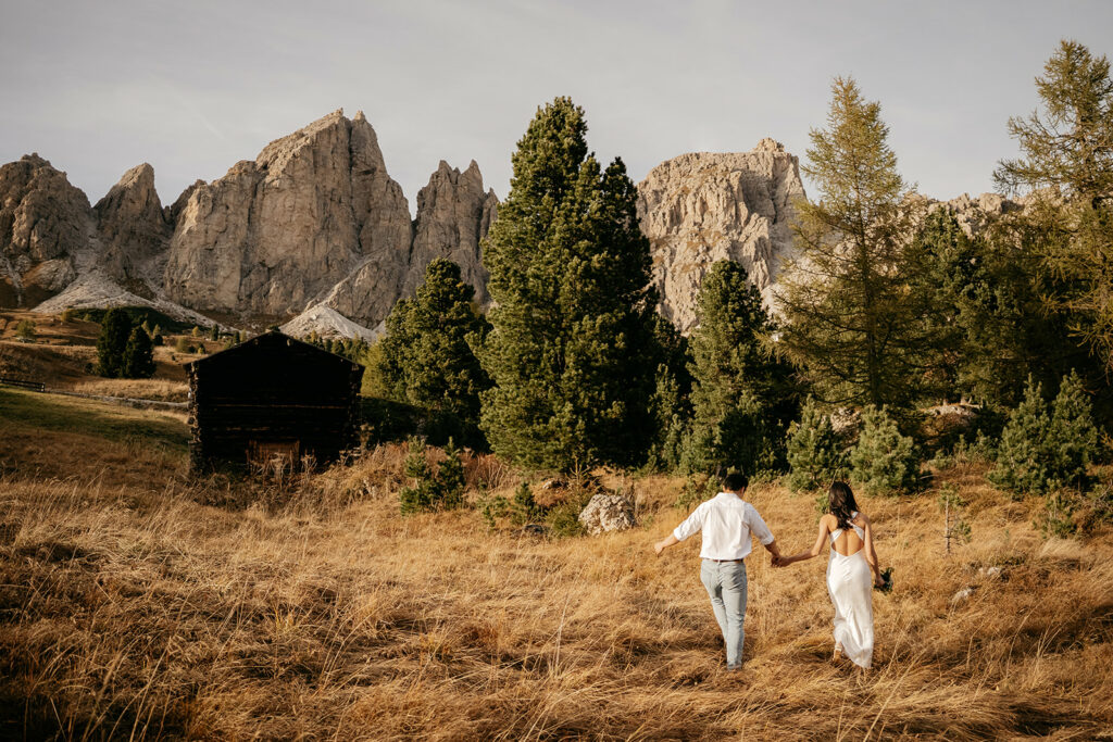 Couple walks through mountain landscape with trees and cabin.