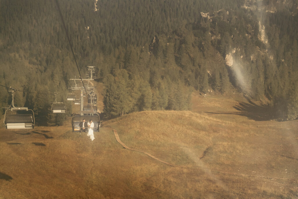 Couple on ski lift over forested mountain slope.