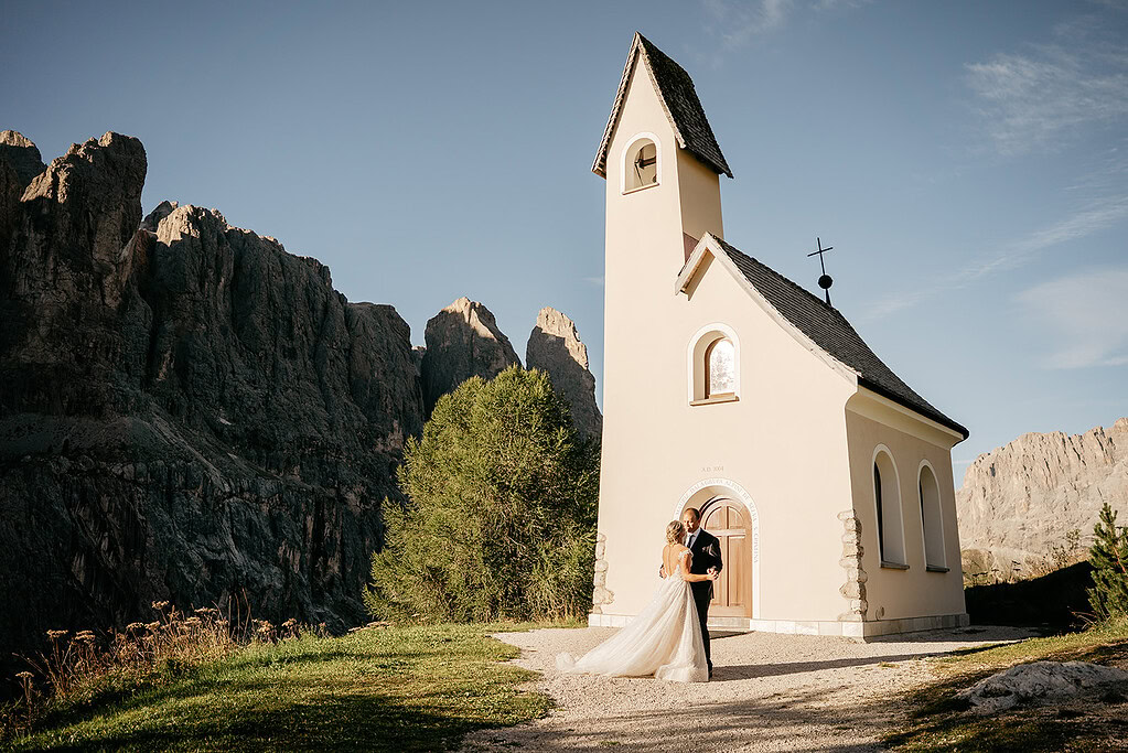 Couple in wedding attire outside small church, mountains behind.
